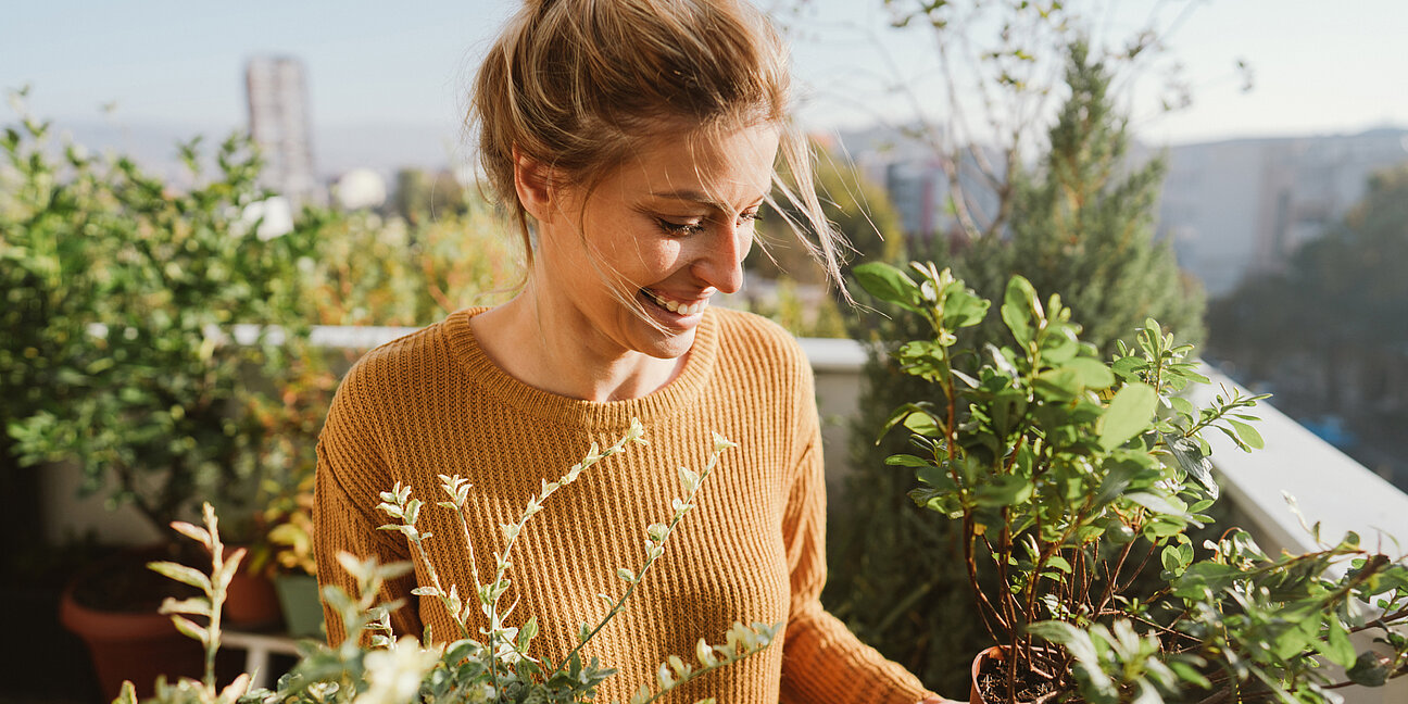 Frau steht lächelnd mit Pflanzen in der Hand auf ihrer Terrasse, passend zum Artikel über Auszahlung beim Neubau.